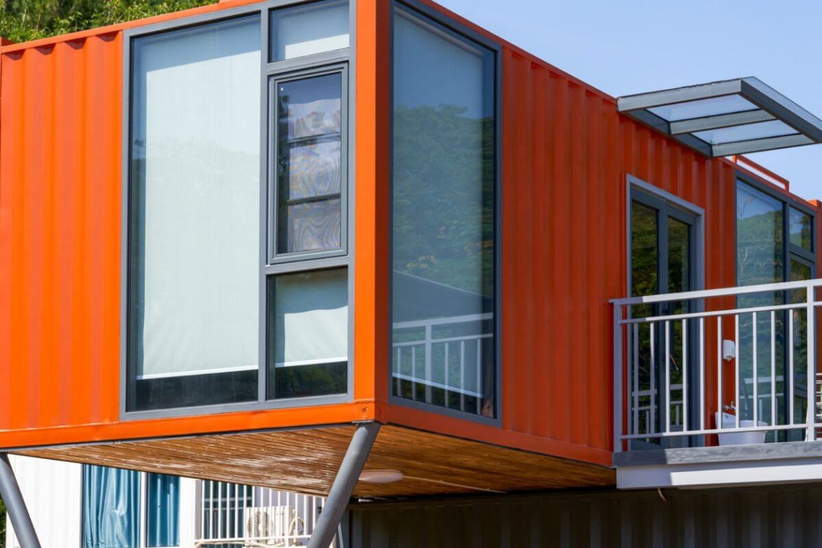 A modern structure in Metro Manila, crafted from an orange shipping container, is elevated on metal supports. It features large glass windows and a small overhanging roof. The surrounding area is lush with greenery and bathed in sunlight, while a railing suggests a balcony's presence.