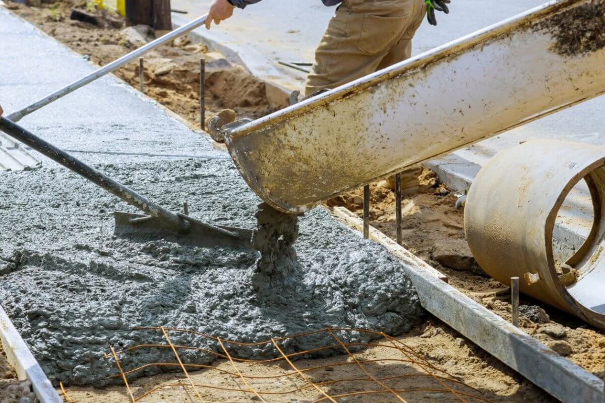 Two workers in Metro Manila pour concrete from a large chute to create a sidewalk. One uses a rake to spread the wet mix evenly. Metal reinforcing bars are visible, and a large concrete pipe lies nearby. The scene is outdoors, amid dirt and construction materials supplied by local concrete mix suppliers.