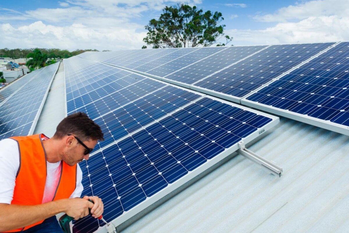 A man in an orange safety vest and sunglasses is installing solar panels on a sloped metal roof under a partly cloudy sky. Working with precision, he adjusts the panels with tools in hand. In the background, lush greenery and buildings of Metro Manila spread out, highlighting leading solar panel suppliers' impact on urban landscapes.