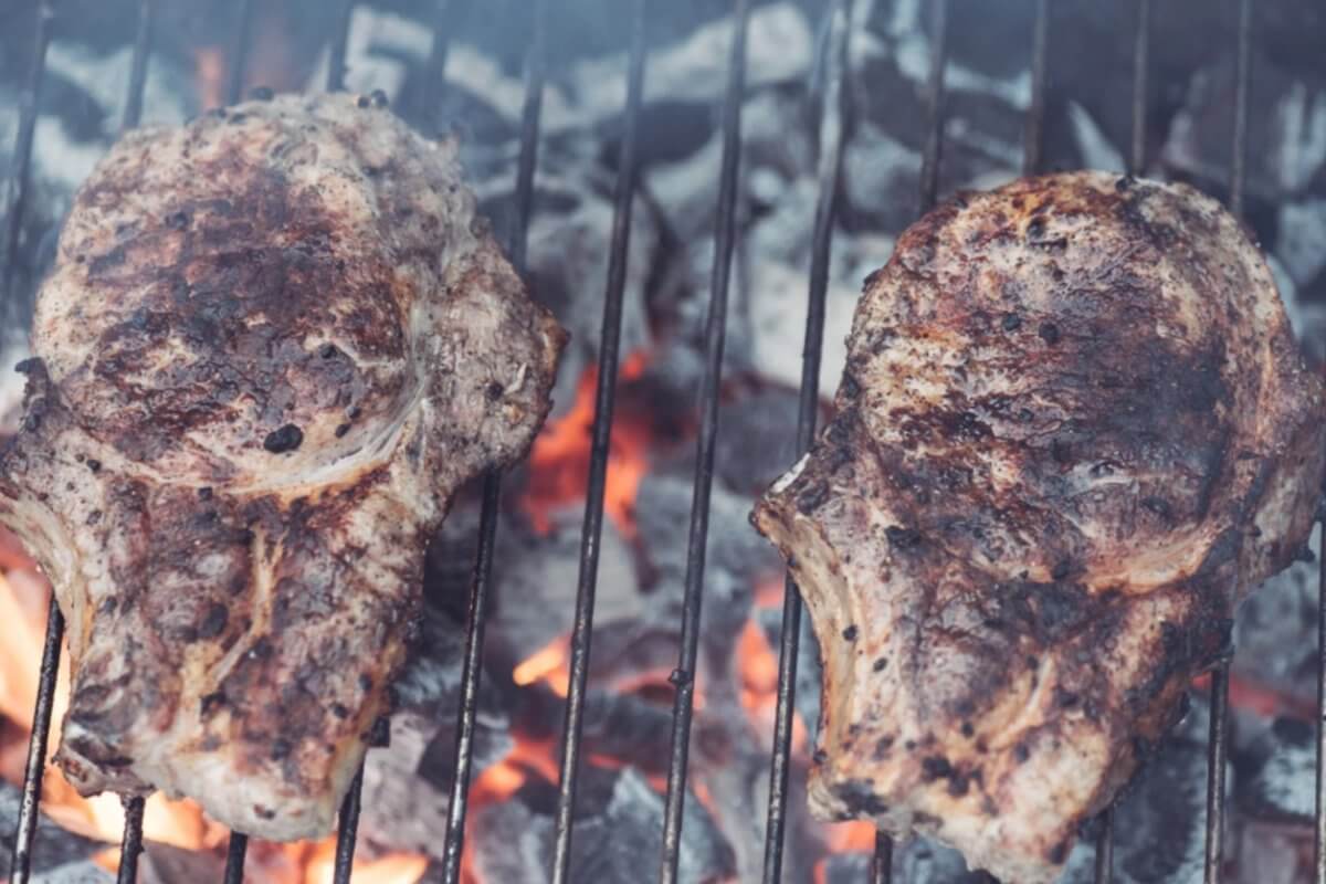 Two seasoned steaks grilling over hot charcoal, with flames and smoke visible between the grill grates, evoke a tantalizing aroma reminiscent of top Binan restaurants.
