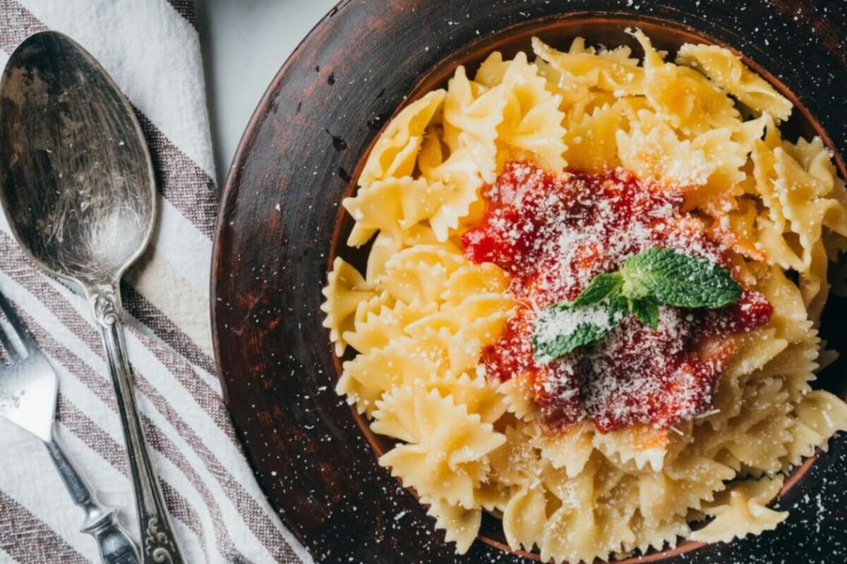 A bowl of farfalle pasta with tomato sauce and mint garnish sits elegantly next to a fork and spoon on a striped cloth, reminiscent of the culinary delights found in Ayala Malls' top restaurants.