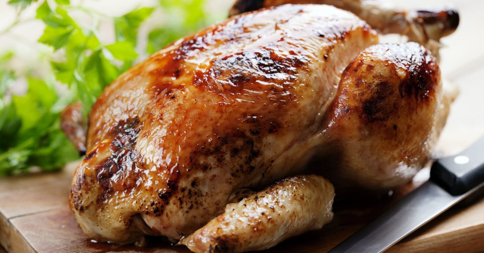 Golden roasted chicken on a wooden board with herbs, reminiscent of the savory delights you'd find at the best restaurants around. A knife is placed beside it, ready to carve into this culinary masterpiece.