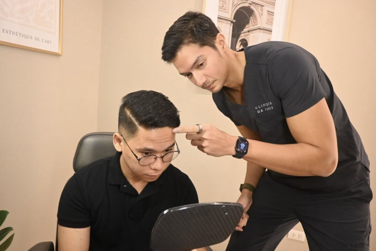 Two men in black shirts, one with a mirror and the other pointing at his head, in a Clinique de Paris consultation room.