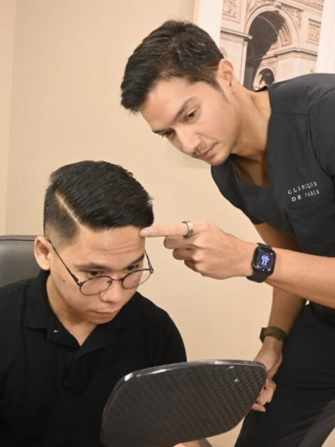 Two men in black shirts, one with a mirror and the other pointing at his head, in a Clinique de Paris consultation room.