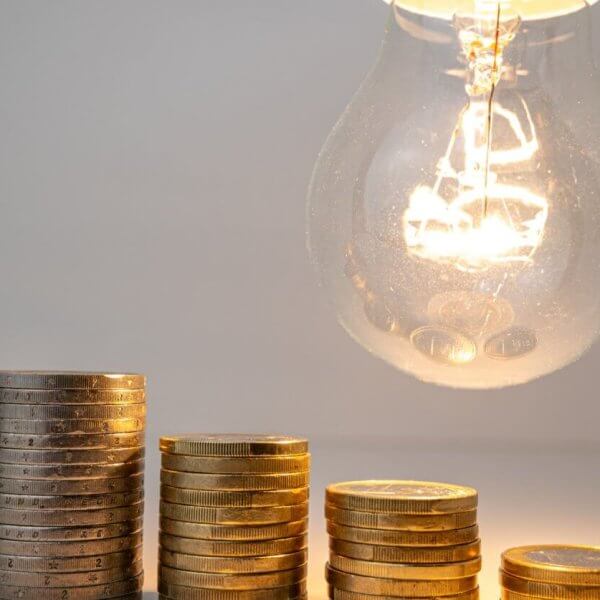 Stacked coins beneath a lit bulb, symbolizing electric consumption and the potential to save with solar.