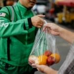 A delivery rider hands over a bag of groceries to a customer at the doorstep — the GrabMart or Pandamart drop-off this guide compares against a personal grocery store trip in the Philippines