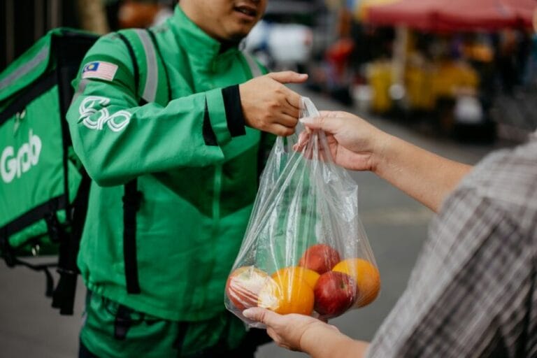 A delivery rider hands over a bag of groceries to a customer at the doorstep — the GrabMart or Pandamart drop-off this guide compares against a personal grocery store trip in the Philippines