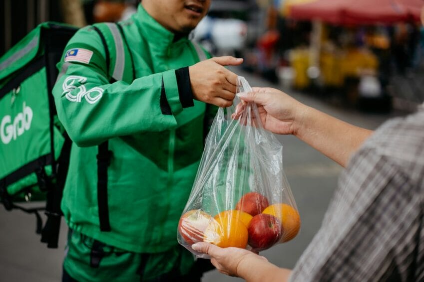 A delivery rider hands over a bag of groceries to a customer at the doorstep — the GrabMart or Pandamart drop-off this guide compares against a personal grocery store trip in the Philippines